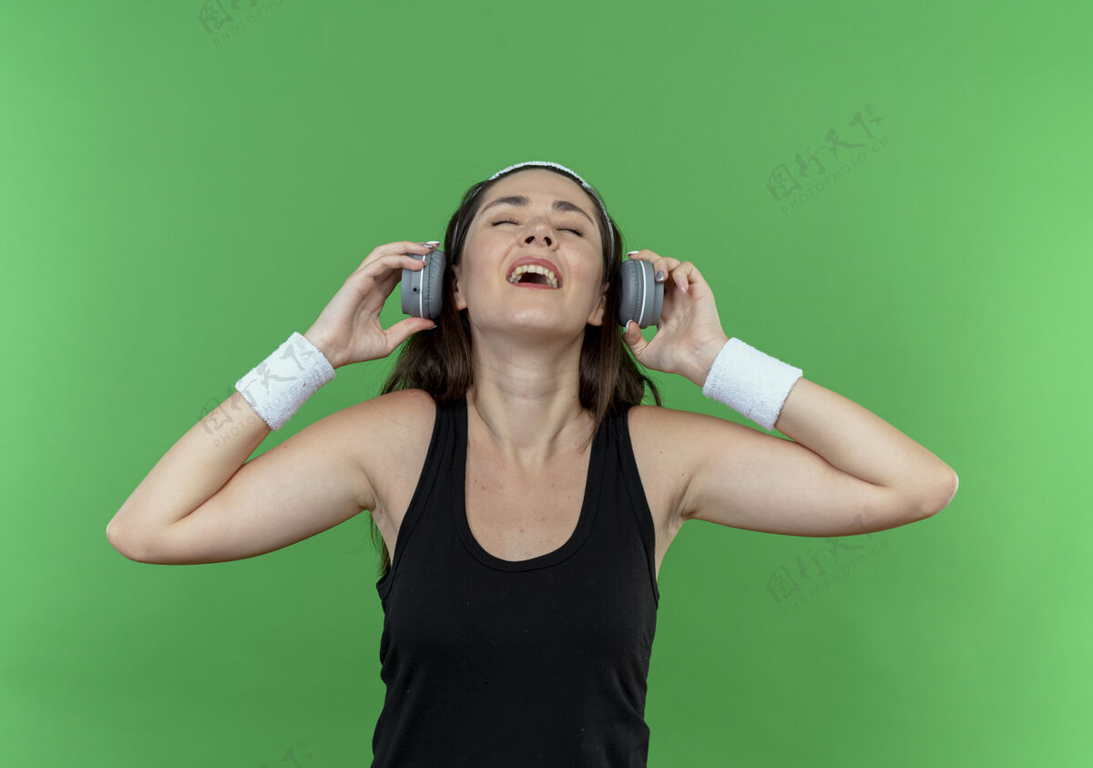 积极戴着耳机戴着头带的年轻健身女士站在绿色的墙上 快乐而积极地欣赏着她最喜爱的音乐头带喜爱音乐
