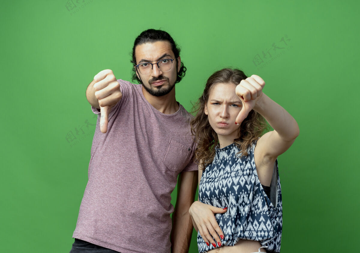 女人年轻的一对男女 不高兴地竖起大拇指站在绿色的墙上夫妇不高兴展示