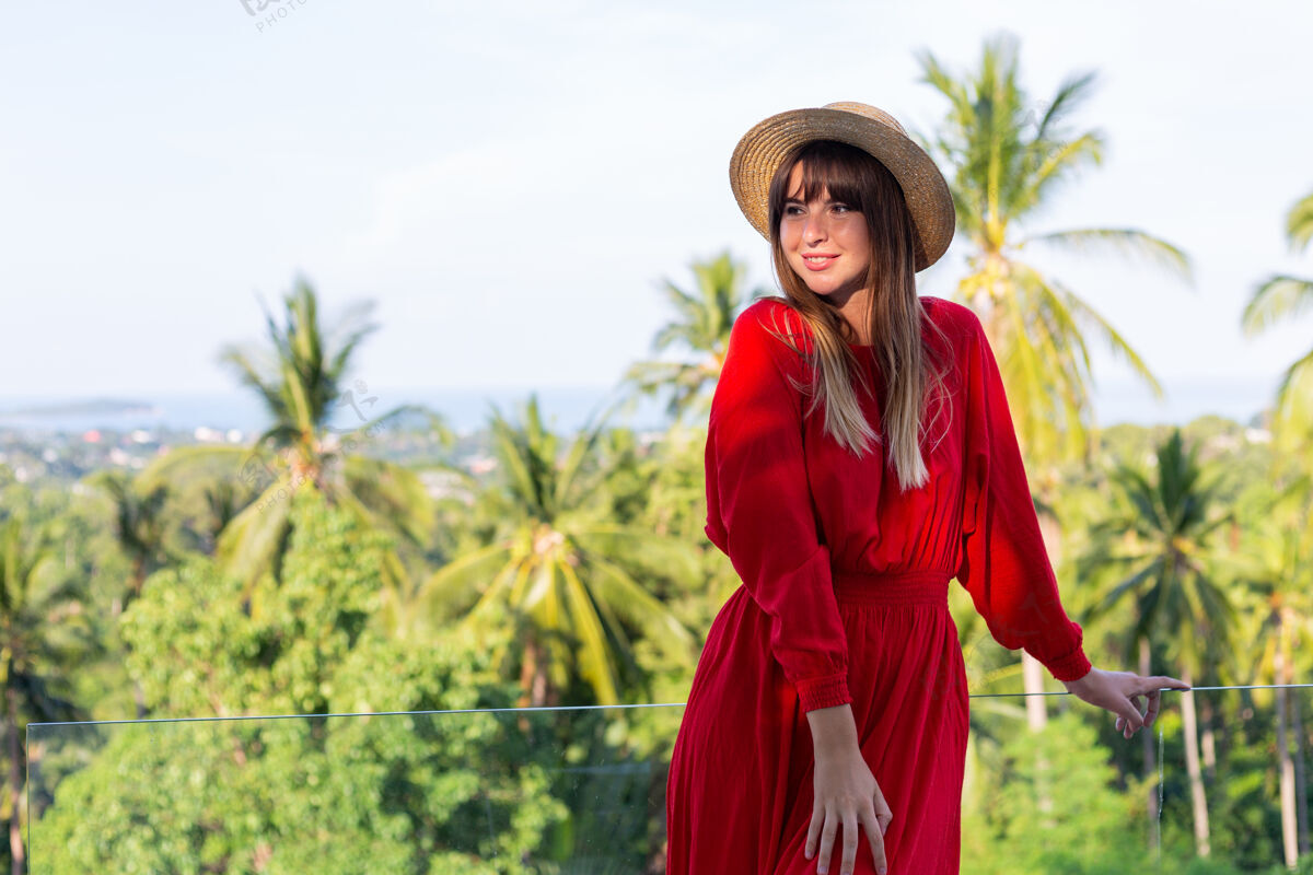 脱毛穿着红色夏装 戴着草帽 在阳台上享受热带海景和棕榈树的快乐女人沙滩优雅帽子
