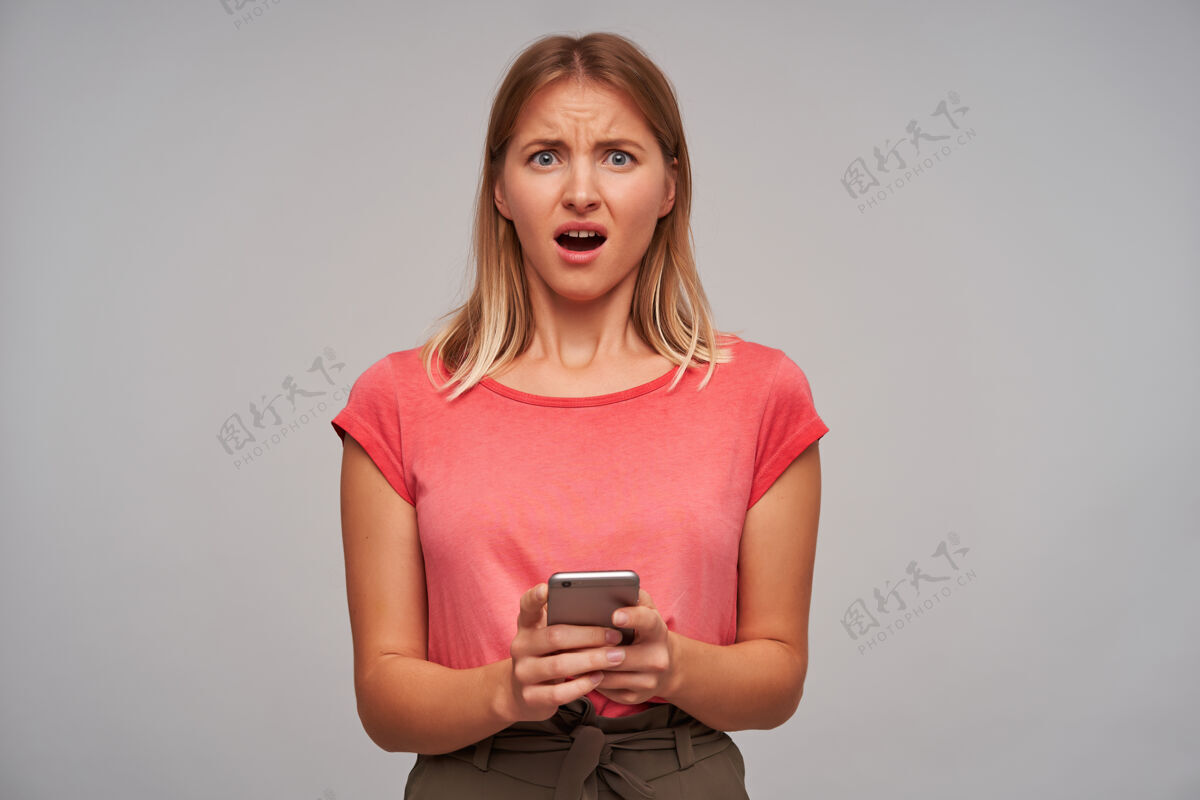20多岁不高兴的 金发的成年女孩的肖像穿着粉色t恤和棕色裙子拿着手机在灰色的墙上收到一个可怕的信息光电话女性