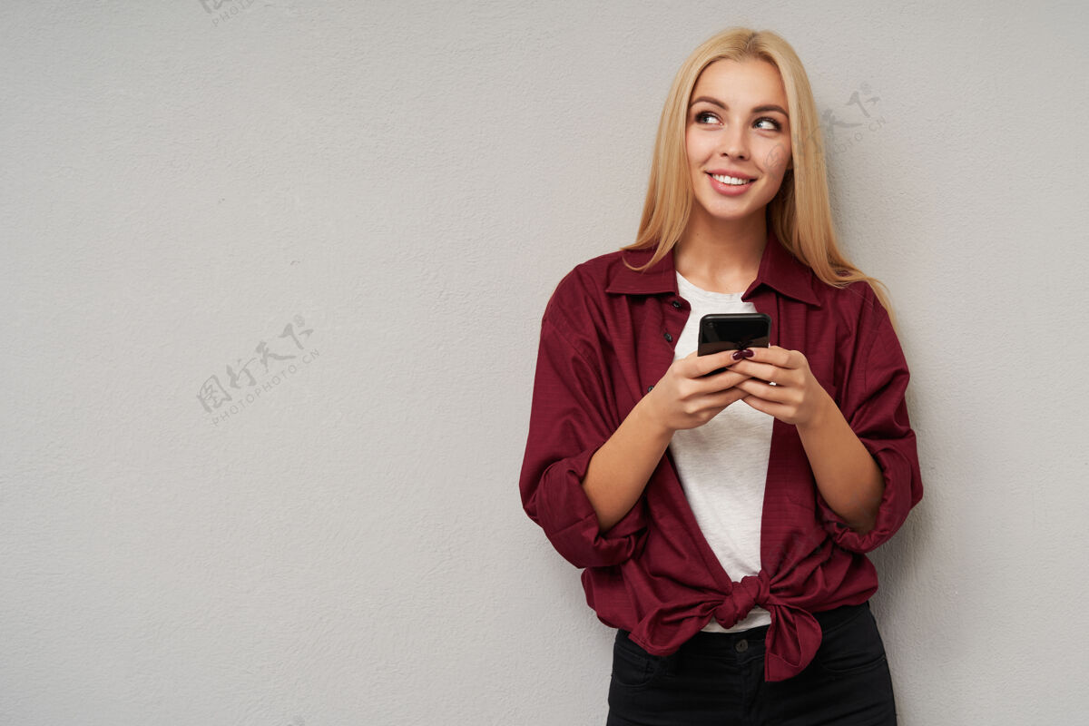 头发迷人的年轻金发女性 留着一头长发 身穿紫红色衬衫和白色t恤 站在浅灰色的旁边 举起手拿着手机 面带温柔的微笑看着一边休闲T恤心情