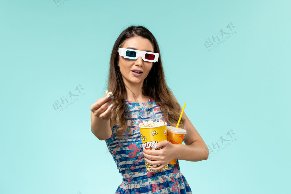 太阳镜正面图年轻女性拿着爆米花包 戴着d墨镜在蓝色表面喝酒前面剧院包装