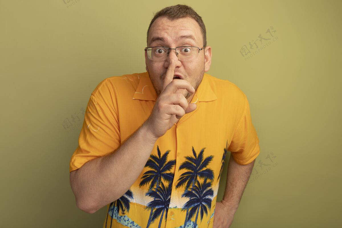 站着戴着眼镜的男人穿着橘色衬衫 手指放在嘴唇上 站在光墙上做着沉默的手势沉默做着手指