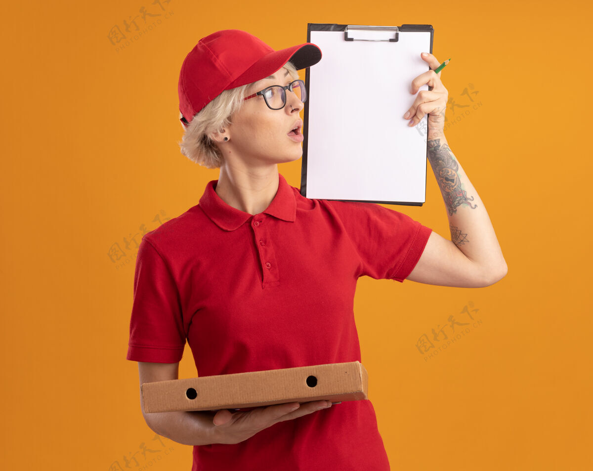 剪贴板身穿红色制服 戴着眼镜 拿着披萨盒的年轻女送货员站在橙色的墙上好奇地看着它展示阴谋披萨