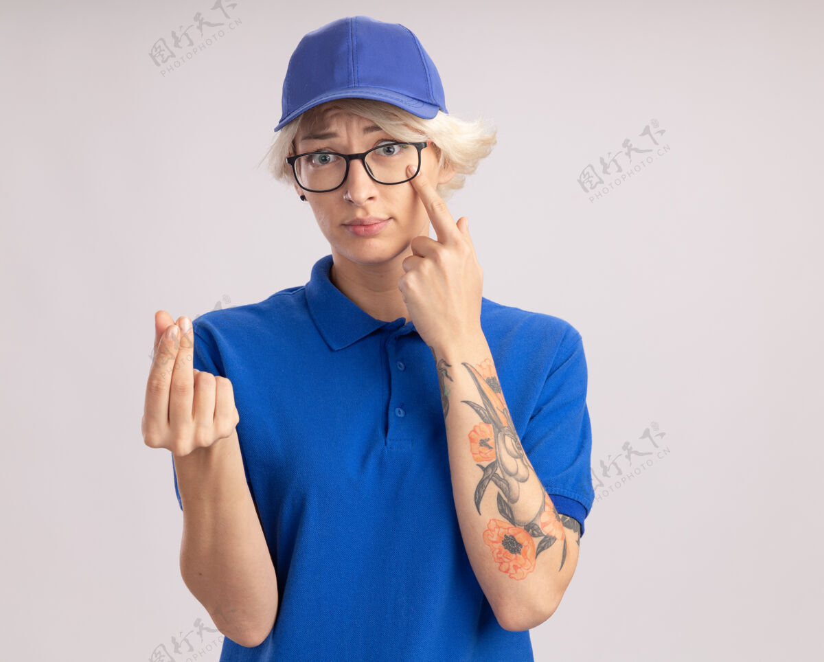 站着身穿蓝色制服 戴着眼镜的年轻送货员 手指指着眼睛 手指搓着手指 站在白墙上做着赚钱的手势指点手指年轻人