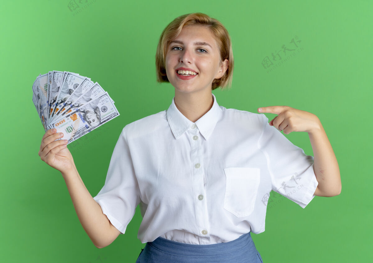 俄罗斯年轻的金发碧眼的俄罗斯女孩抱着钱 指着隔离在绿地上的复制空间持有年轻金钱