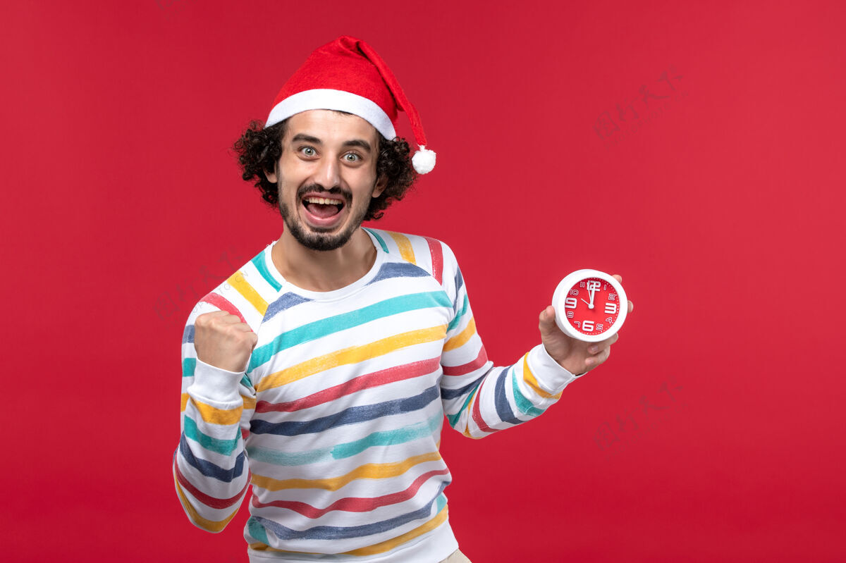 男性正面图年轻男子高高兴兴地拿着红墙上的时钟新年假期红男风景帽子举行