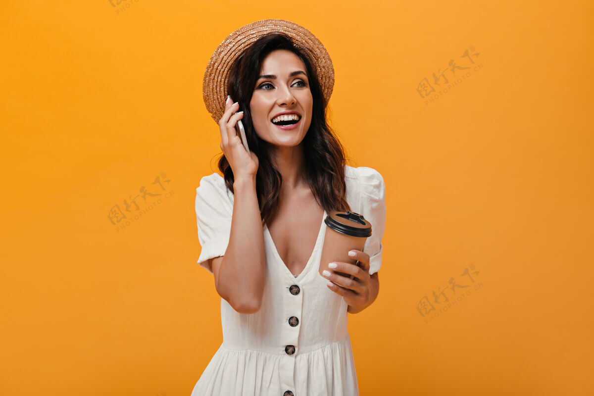 室内戴草帽的快乐女孩正在讲电话 手里拿着一杯咖啡穿着鲜艳衣服的漂亮女士在孤立的背景下拿着智能手机和茶裙子苍白衬衫