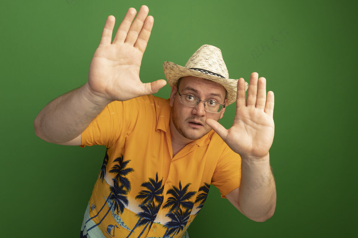 防御一个戴着眼镜的男人穿着橙色衬衫 戴着夏令帽 站在绿色的墙壁上做着防卫的手势 手被吓坏了眼镜帽子夏天
