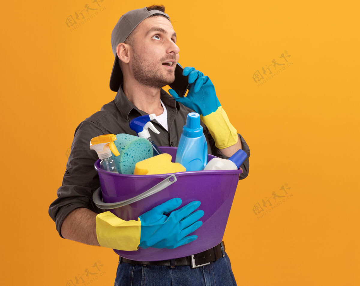 自信年轻人穿着休闲服 戴着橡胶手套 手里拿着水桶 手里拿着清洁工具 站在橘黄色的墙上 一边微笑着自信地说着手机工具水桶电话