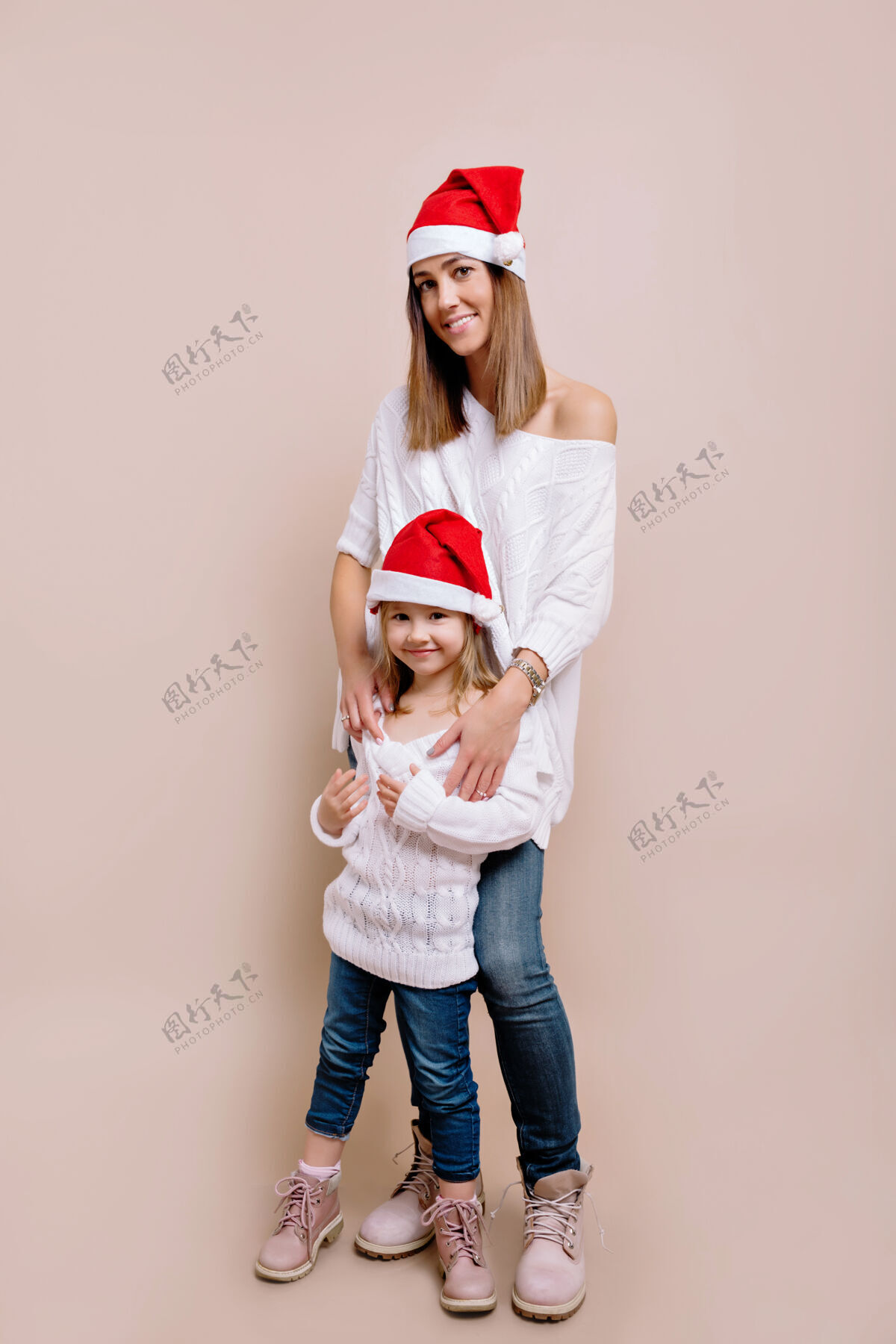 家庭工作室的肖像画 年轻漂亮的女人和她的小女儿穿着白色毛衣和圣诞帽快乐冬天年轻