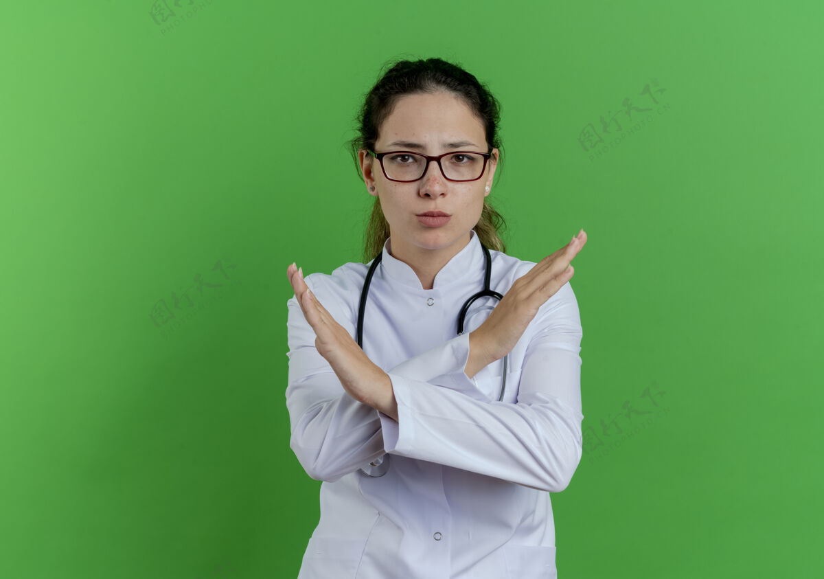 表情严格的年轻女医生穿着医用长袍 戴着听诊器和眼镜 做着没有手势的隔离在绿色的墙上 留着复印空间年轻眼镜严格