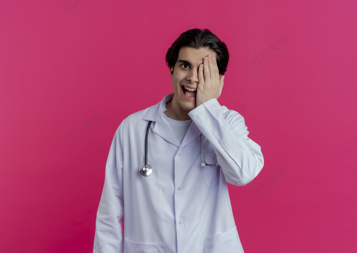 医生令人印象深刻的年轻男医生穿着医用长袍和听诊器覆盖半张脸隔离在粉红色的墙壁与复制空间男性半身戴
