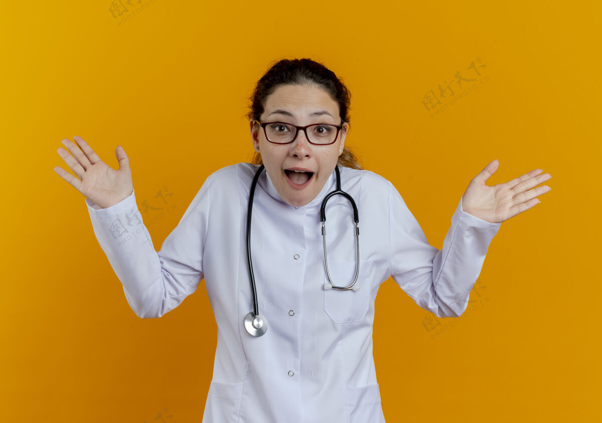 惊喜年轻的女医生穿着医用长袍 戴着听诊器 戴着眼镜 双手被隔离在橙色的墙上手长袍医生