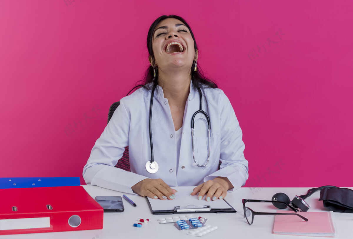 女笑着的年轻女医生穿着医用长袍和听诊器坐在办公桌旁 手里拿着医疗工具 双手放在办公桌上 闭着眼睛隔离在粉红色的墙上年轻医生医疗