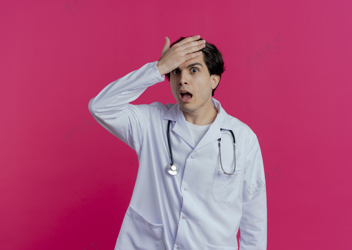 男性印象深刻的年轻男医生穿着医用长袍和听诊器把手放在额头上做现在我记得我忘了什么 隔离在粉红色的墙上与复制空间印象医生额头