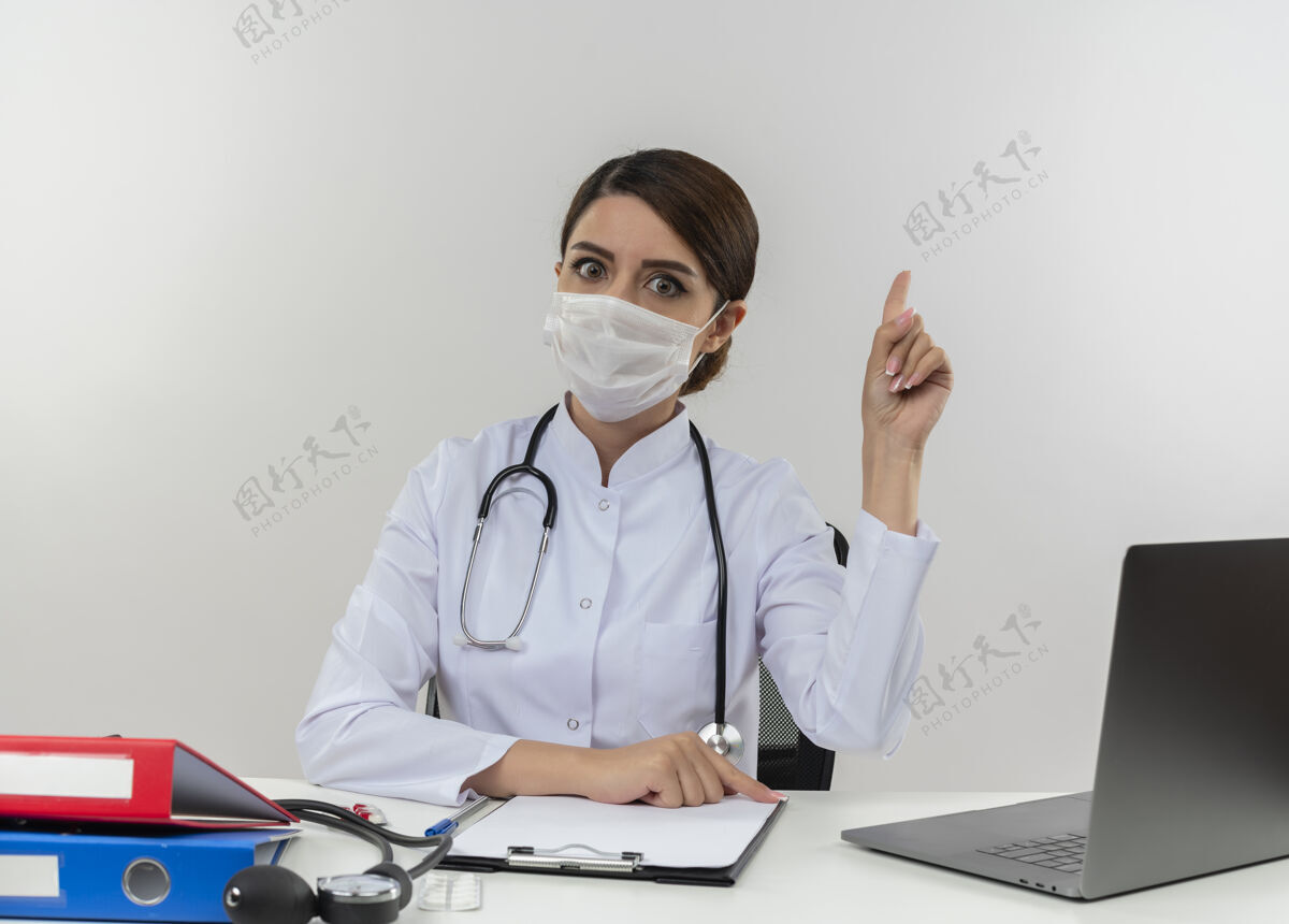 笔记本电脑年轻的女医生穿着医用长袍 戴着听诊器和医用面罩 坐在办公桌旁 手持医疗工具和笔记本电脑 孤零零地站在白色的墙上年轻人穿着坐着
