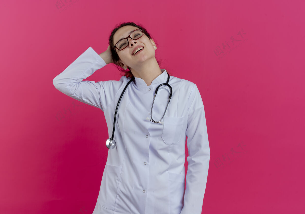 医疗很高兴年轻的女医生穿着医用长袍 戴着听诊器和眼镜 手放在脑后 闭着眼睛 隔离在粉红色的墙上 留着复印空间眼镜女封闭