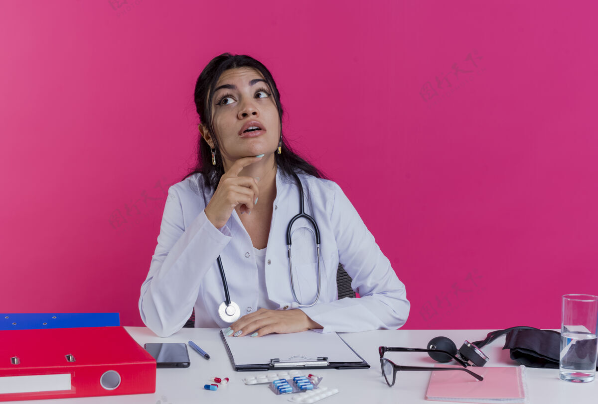 听诊器年轻的女医生穿着医用长袍 手持听诊器坐在办公桌旁 手持医疗工具 手放在办公桌上 一边看着一边 一边抚摸着粉红色墙上的下巴放工具穿