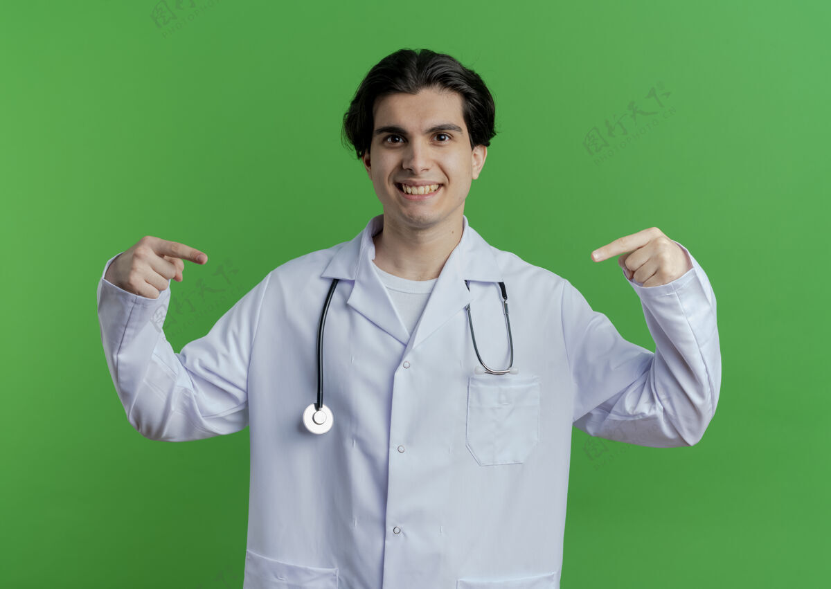 微笑面带微笑的年轻男医生 穿着医用长袍 手持听诊器 指着他面前被隔离在绿色墙上的东西前面医疗听诊器