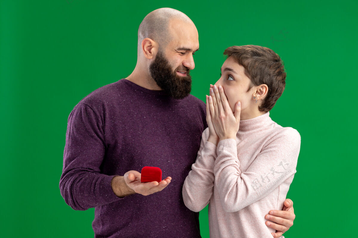 女朋友一对穿着休闲装的年轻情侣站在绿色的背景下 带着红盒子里的订婚戒指 幸福地向他惊奇的女友求婚惊人休闲年轻