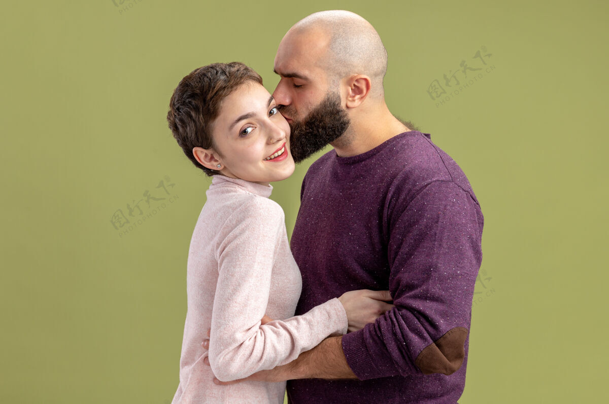 立场一对穿着休闲装的年轻夫妇一个留着短头发和胡须的女人一个热恋中的男人站在绿色的墙上亲吻他的女友庆祝情人节休闲白天胡须