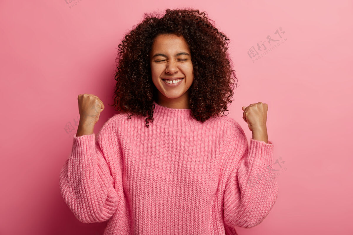 女人快乐的黑皮肤少年举起紧握的拳头 庆祝胜利和成功 获得理想的愿望 笑容灿烂 穿着针织套头衫 隔离在粉红色的墙上年轻是服装