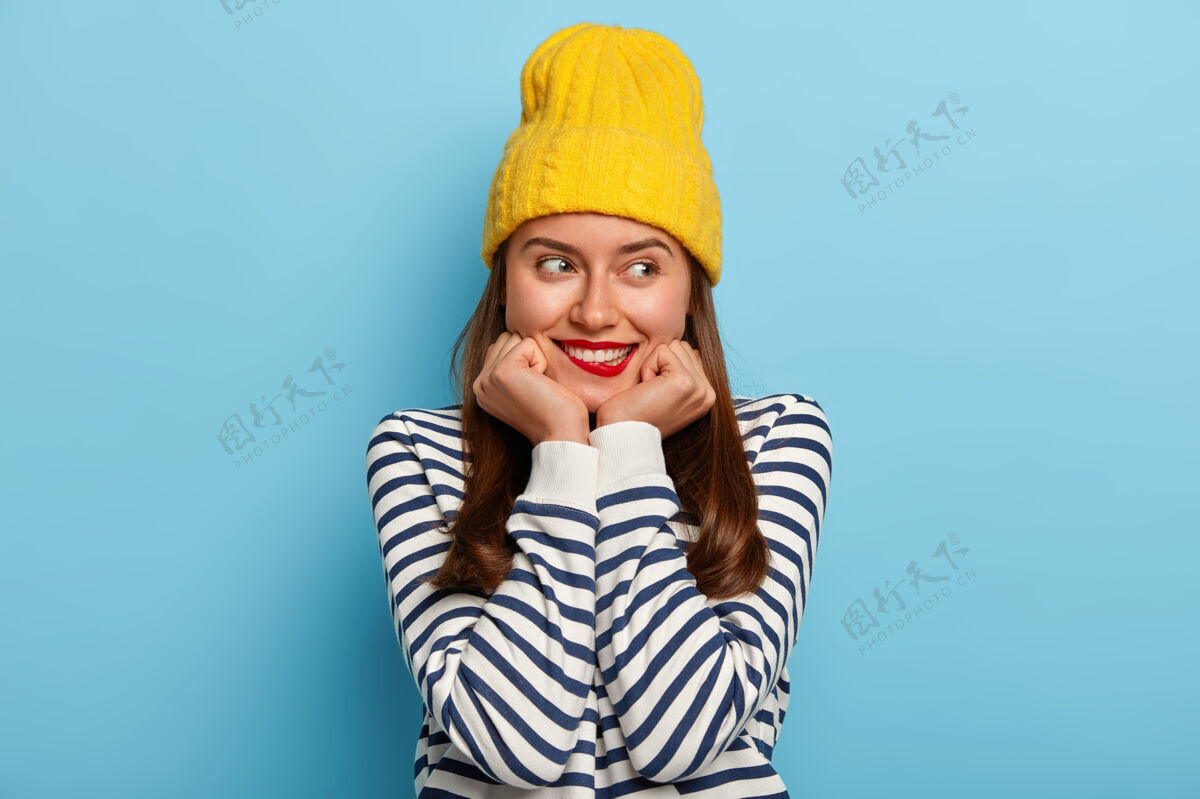 水平美丽的黑发千禧一代女人双手放在下巴下 咬着嘴唇 有着满意的表情 戴着黄色帽子和条纹套头衫黑发冬天欧洲人