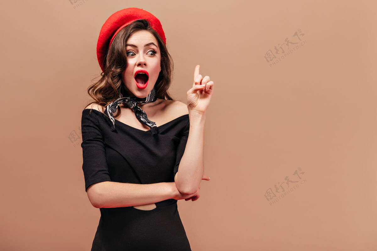 米色背景穿黑衣服 戴红帽子 戴红围巾的年轻女士想出了一个新主意 食指朝上伸了出来脸长卷发