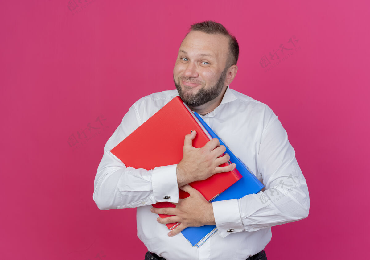 男士高兴的胡须男子穿着白衬衫拿着文件夹微笑愉快地超过粉红色穿上微笑衬衫