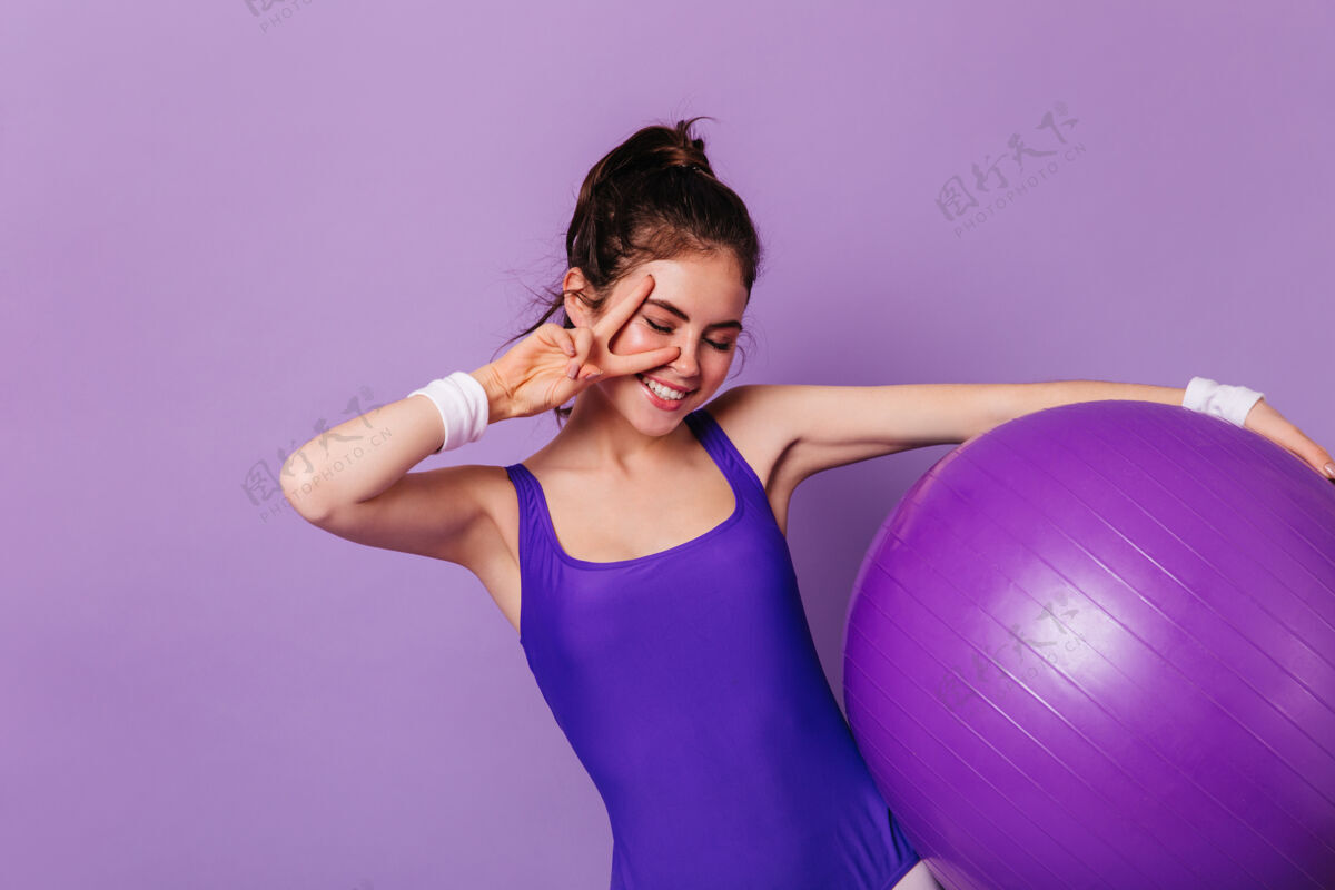 紧身裤苗条的体操女子手持健身球 在紫色的墙上显示和平标志健康健美操时尚