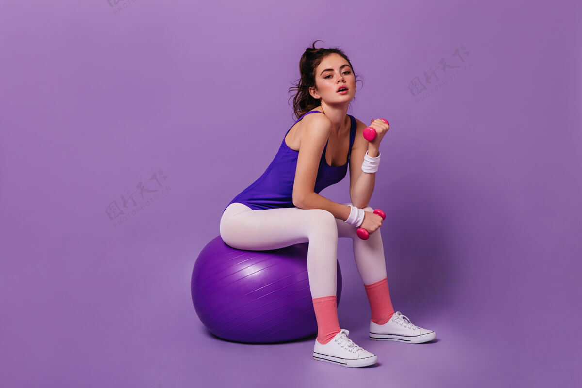 卷发穿着运动紧身衣和粉色袜子的年轻女子坐在fitball上 在紫色的墙上和哑铃握手锻炼运动身材
