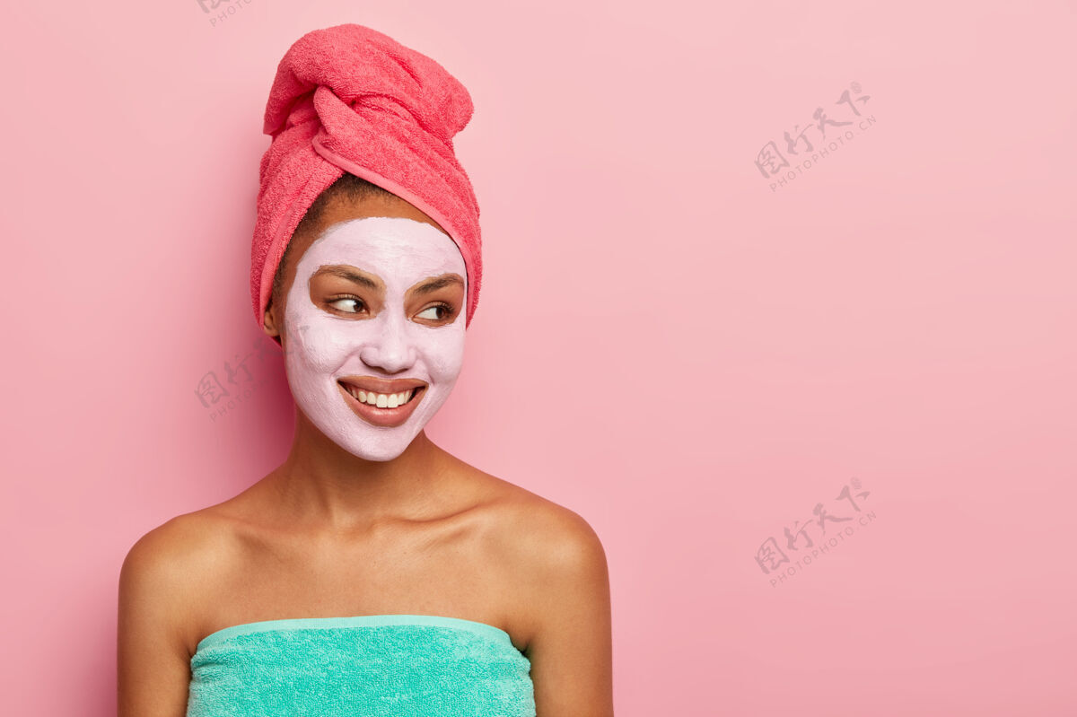 健康裹着毛巾的清爽女人 涂上减少脸上细纹的泥质面膜 开心地看着一旁 对着粉色的墙壁摆姿势女人魅力皮肤护理