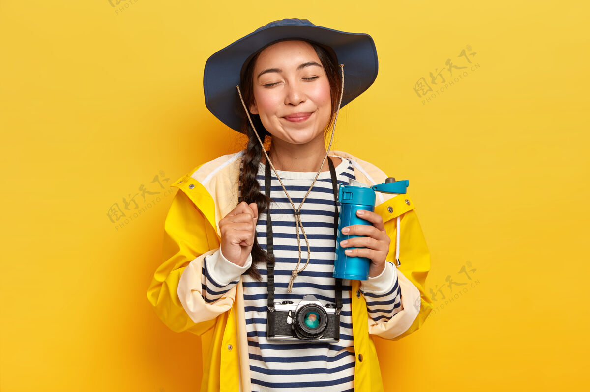 咖啡满意的亚裔女性旅行者 戴着帽子 条纹套头衫和雨衣 脖子上挂着复古相机 手里拿着一瓶热饮 隔着黄色的墙度假年轻发现