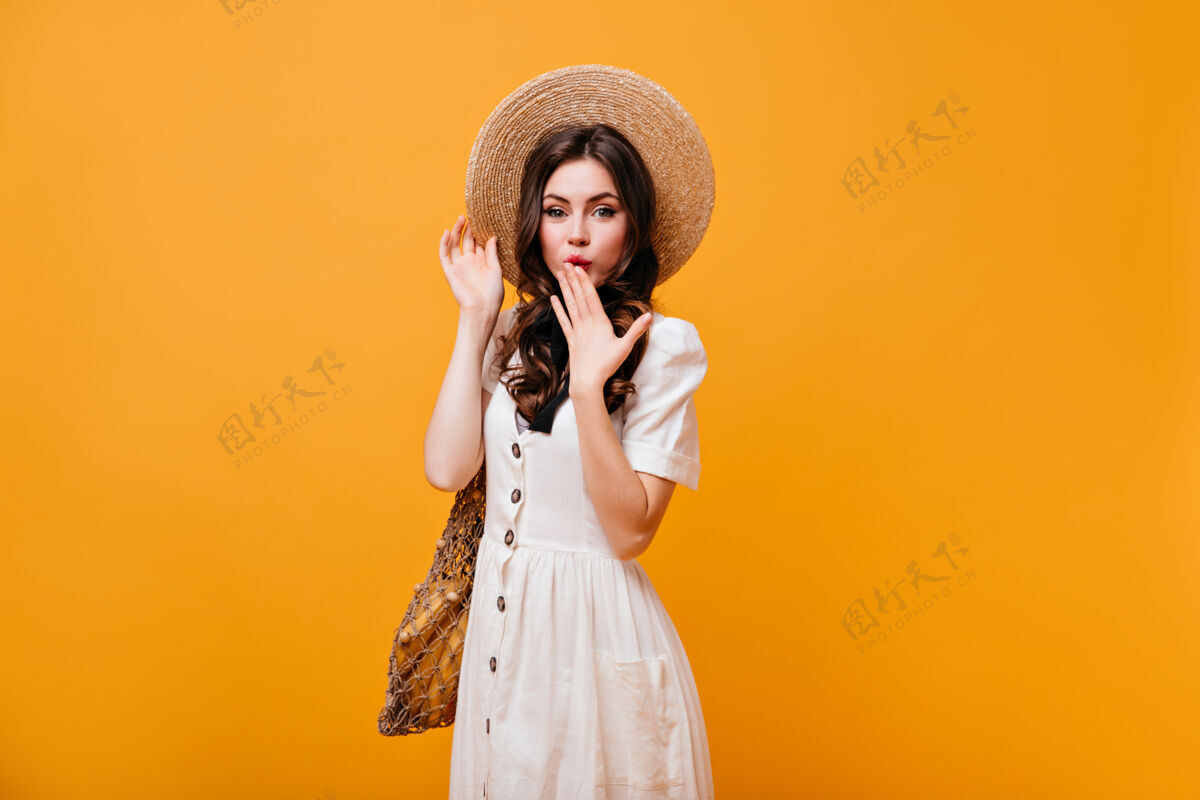 肖像绿眼睛的女士用手捂住嘴戴草帽和白色太阳裙的女士拿着橙色背景的购物袋头发脸年轻人