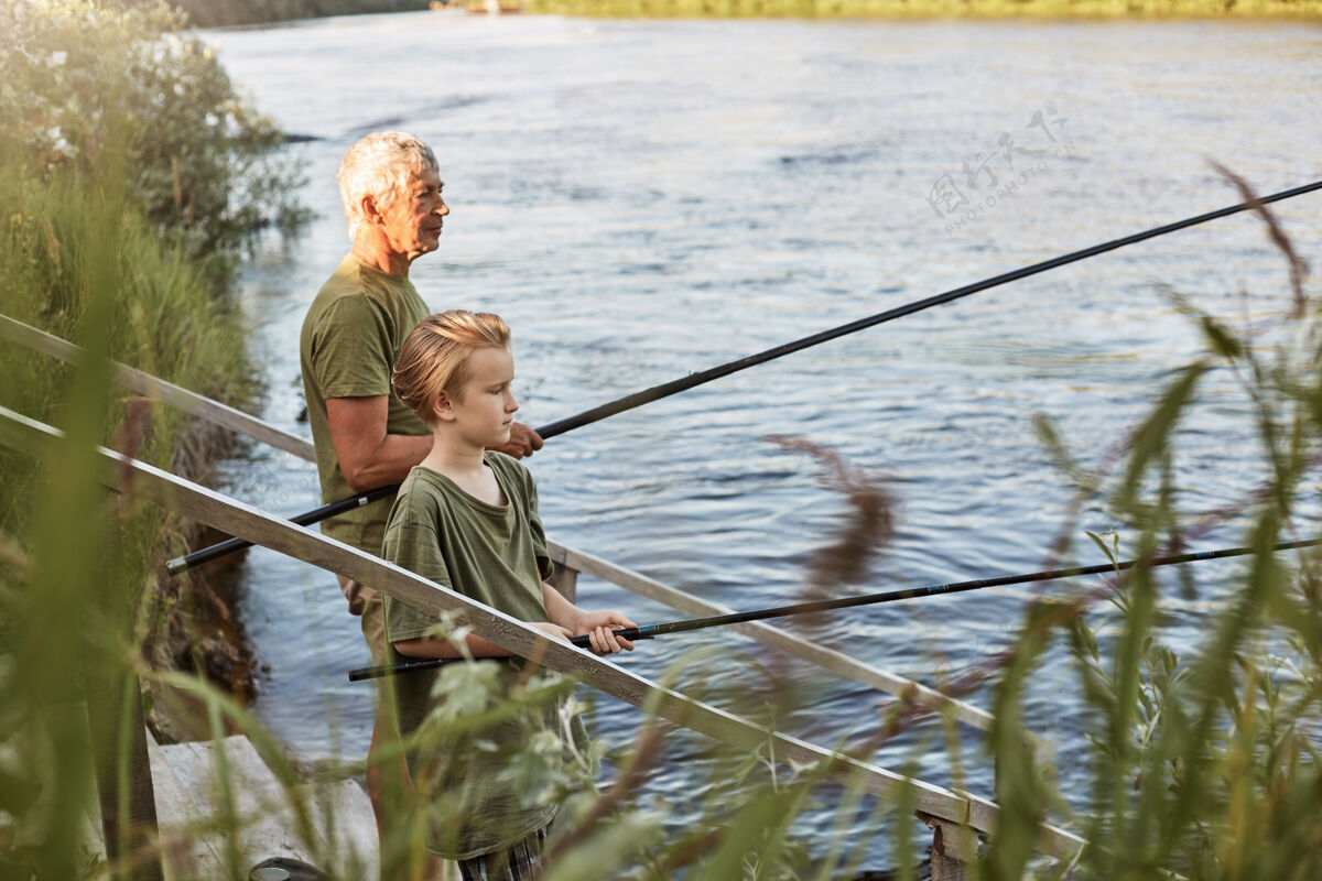 钓鱼欧洲灰白头发的成熟父亲带着儿子在湖边或河边钓鱼 站在水边 手里拿着鱼竿 穿着随意 享受爱好和自然小父亲幼儿