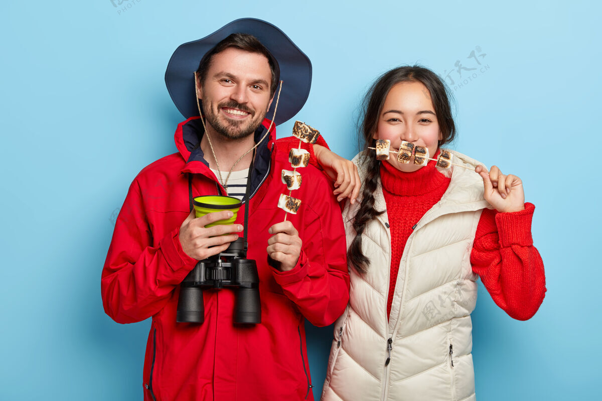 微笑情侣们一起野餐 享受与大自然的独处 吃烤棉花糖喝咖啡 心情愉快 对着蓝色的墙壁摆姿势饮料休闲饮食