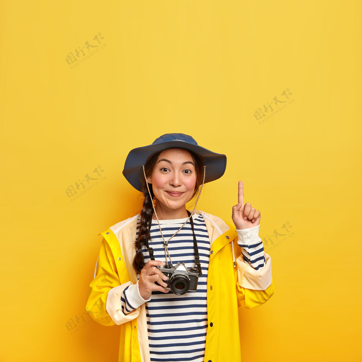 女性美丽的黑发女性 亚洲外貌 在徒步旅行中用复古相机拍照 穿着条纹套头衫 帽子和雨衣 食指指向上方 隔离在黄色的墙壁上人指着高兴