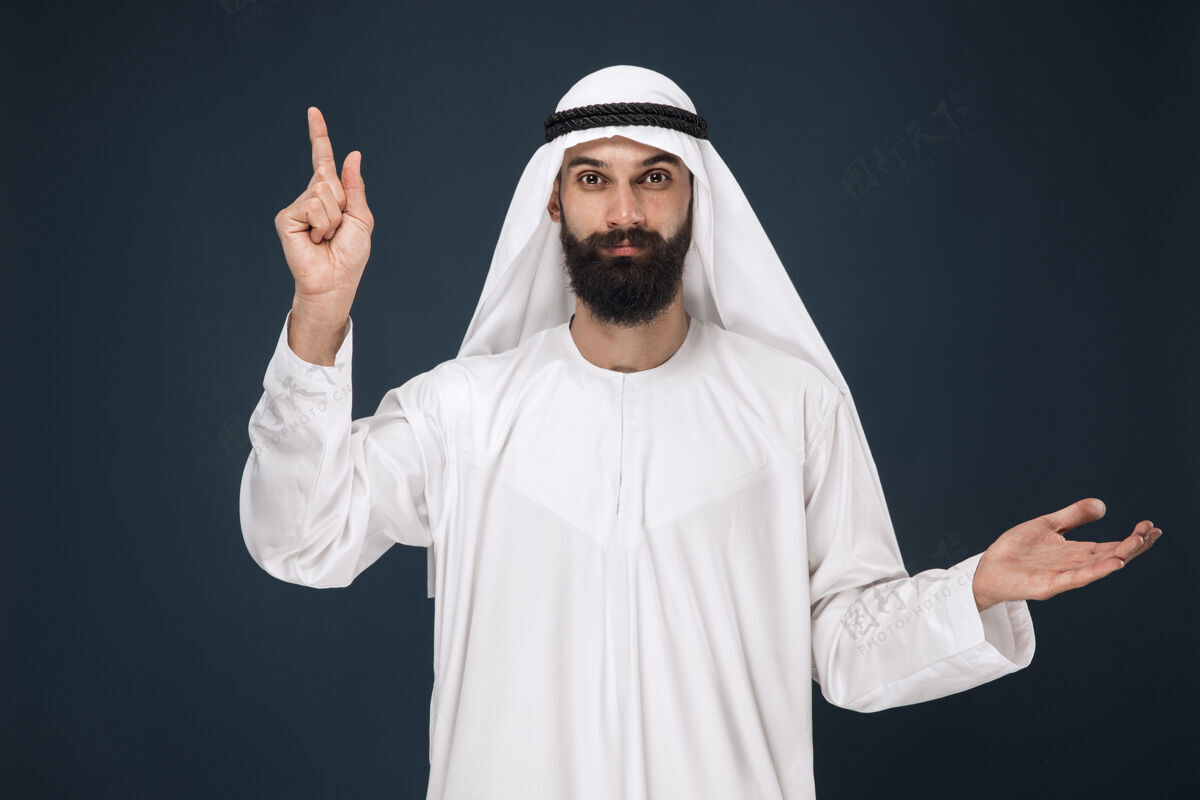 男性深蓝色墙上的阿拉伯沙特男子半身像年轻的男模微笑着指着商业 金融 面部表情 人类情感 技术的概念衣服反应发呆