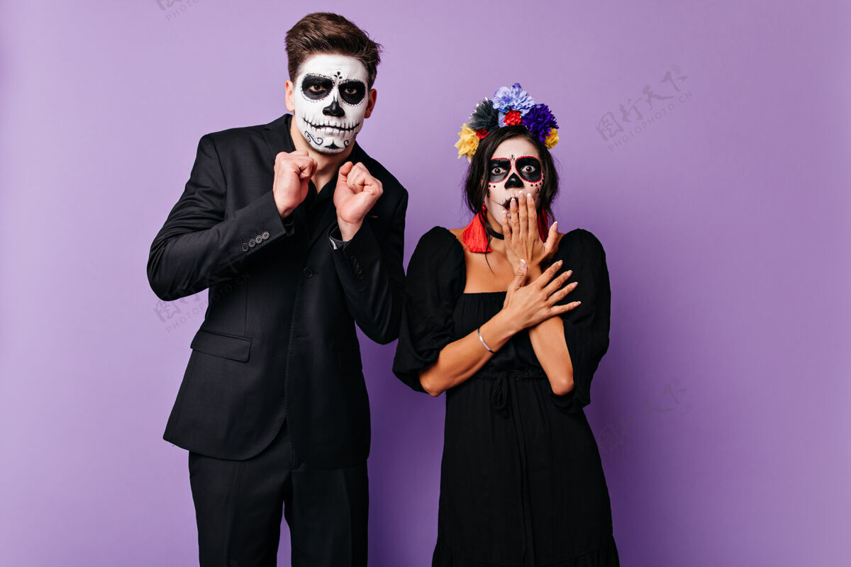 幻想一对戴着骷髅状面具的夫妇在紫色的墙上惊恐地摆着姿势照片上是一个穿着黑色西装的男人和一个穿着深色裙子 带着明亮口音的女孩墨西哥嘉年华传统
