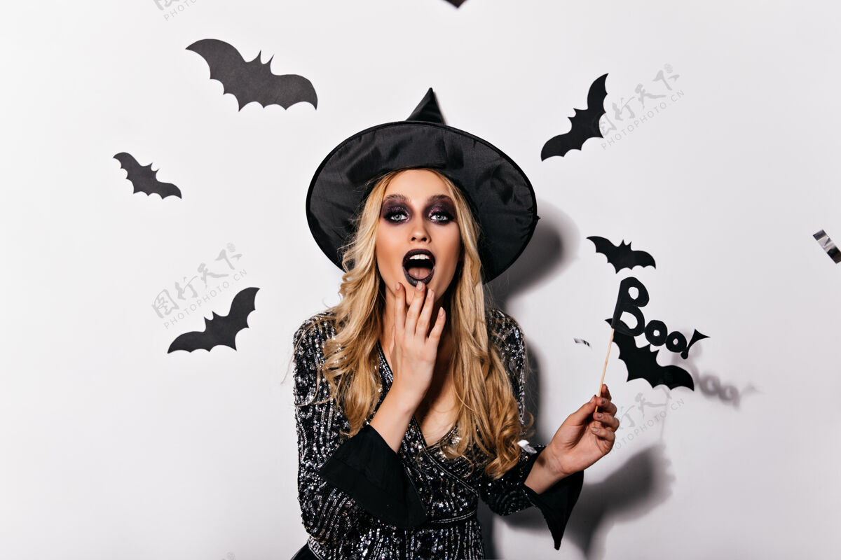 嘉年华一个穿着女巫服装的感兴趣的金发女人在白墙上开玩笑地摆姿势被蝙蝠包围的女吸血鬼万圣节蝙蝠神秘
