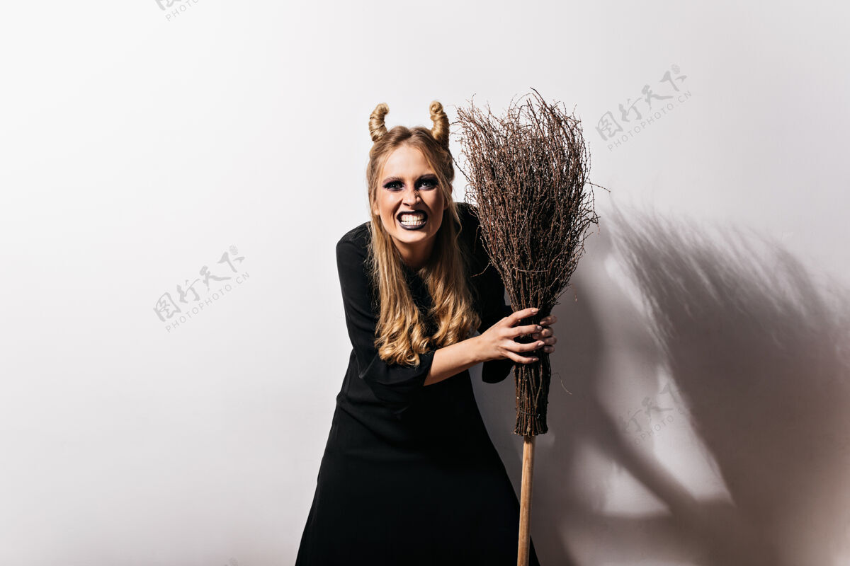女巫快乐的女巫拿着扫帚 滑稽的发型室内拍摄的穿着万圣节服装站在白墙上的兴奋女孩金发邪恶神秘