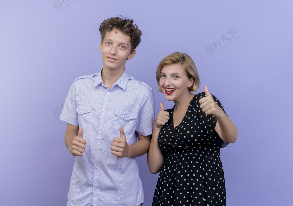 看年轻漂亮的夫妇站在蓝色的墙上微笑着竖起大拇指男人微笑女人