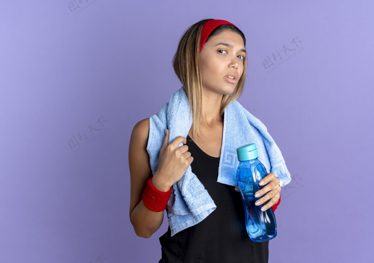 毛巾身穿黑色运动服 头戴红色头巾 脖子上围着毛巾的年轻健身女孩 手里拿着一瓶水站在蓝色的墙上抱女孩脖子