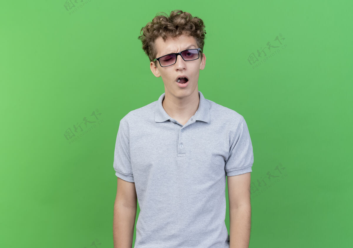 看一个戴着黑眼镜 穿着灰色马球衫 满脸怀疑的年轻人站在绿色的墙上站表情怀疑