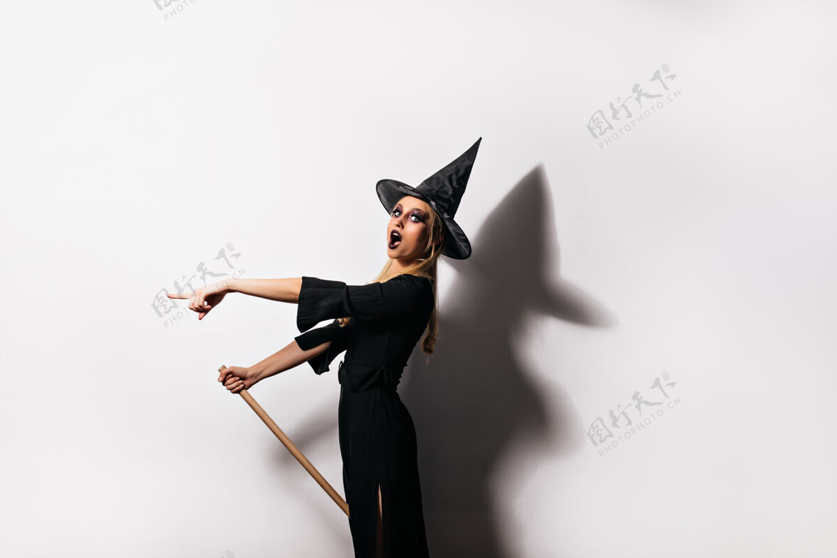 魔术师穿着女巫服装的苗条震惊的女人摆姿势室内拍摄的迷人的黑帽子巫师金发扫帚帽子