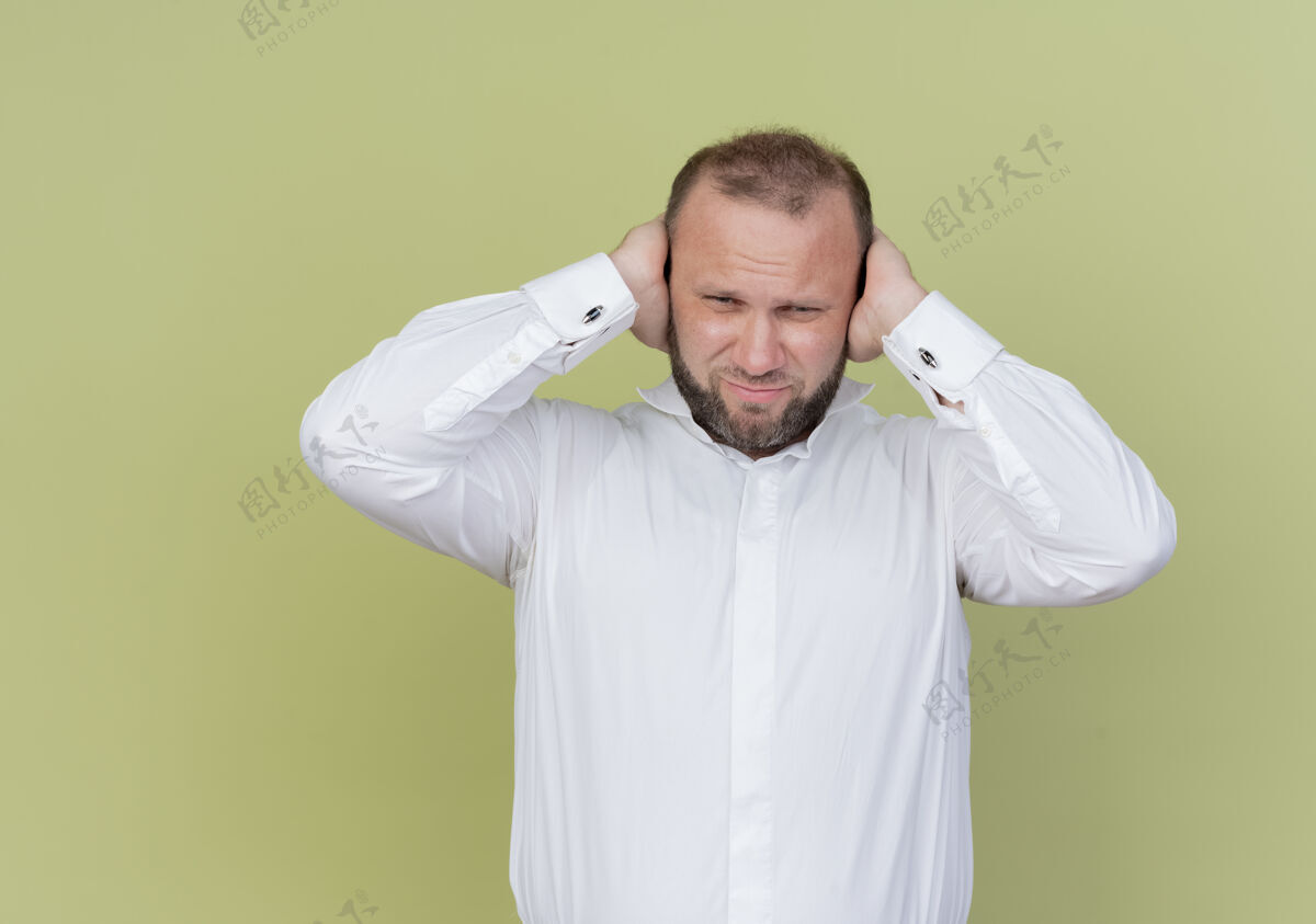 穿留着胡子的男人穿着白衬衫 看起来很困惑 闭着耳朵 双手站在轻墙上站看迷茫
