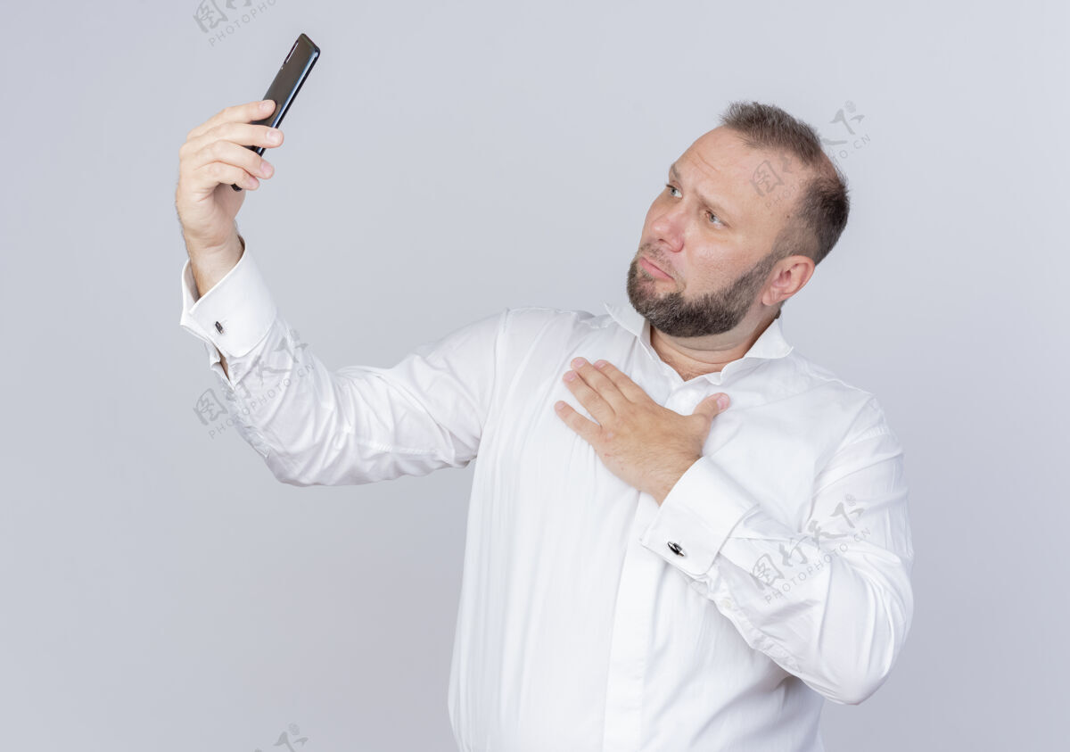 站着留着胡子的男人穿着白衬衫拿着智能手机打视频电话感激不尽手放在胸前站在白墙上视频电话男人