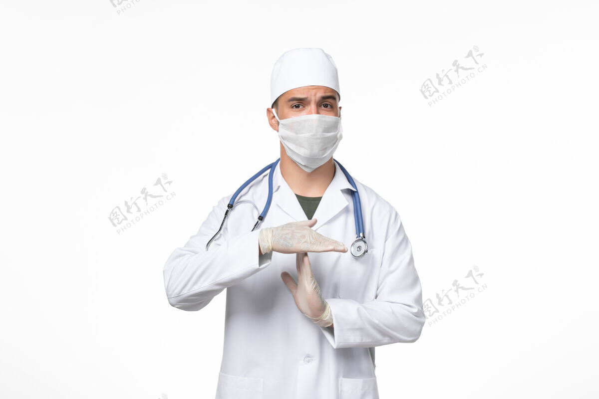 视图正面图：男医生穿着医疗服 戴着口罩 对抗白墙上的冠状病毒病医学大流行病毒实验室外套听诊器医生