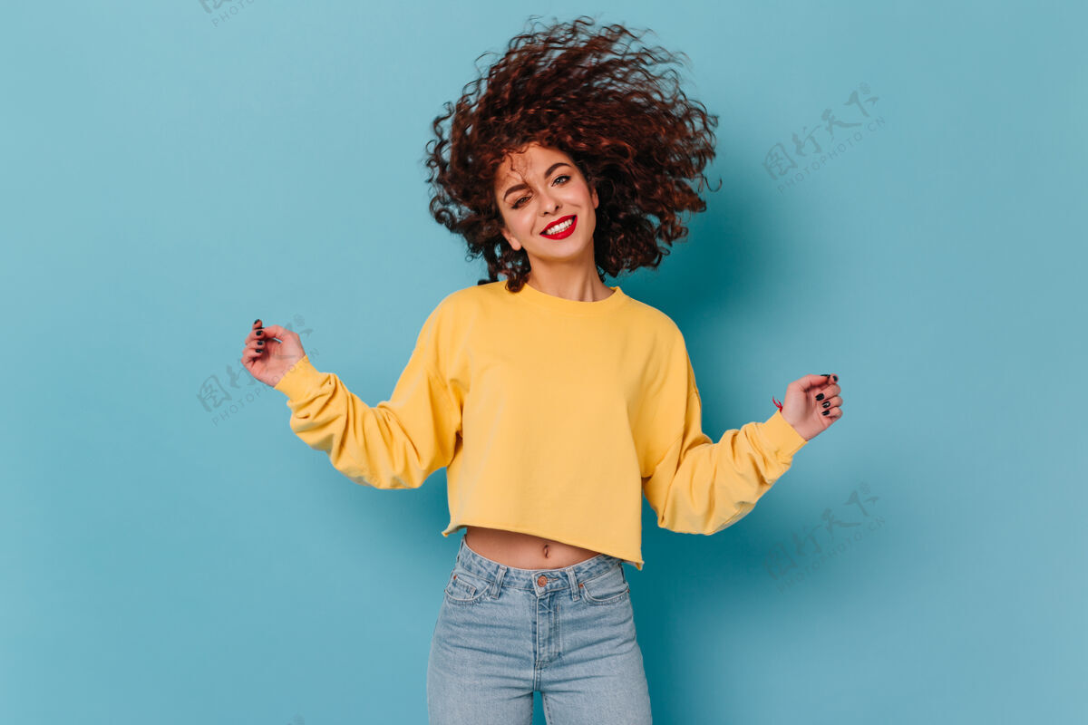 头发微笑的卷发女人 红唇 穿着黄色毛衣和紧身牛仔裤 在与世隔绝的空间里玩头发卷曲高兴红色唇膏